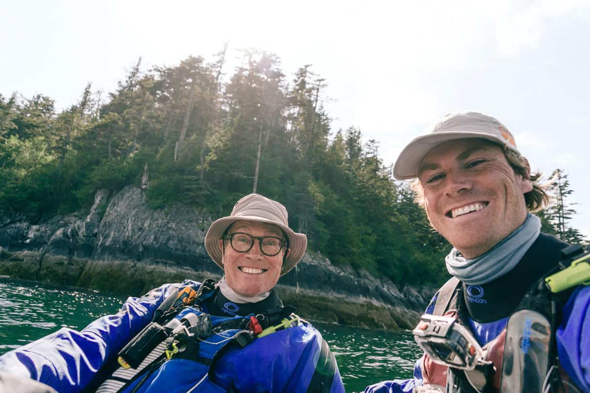 Oli and his dad smile while floating in front of a rocky coast on their sea kayaks