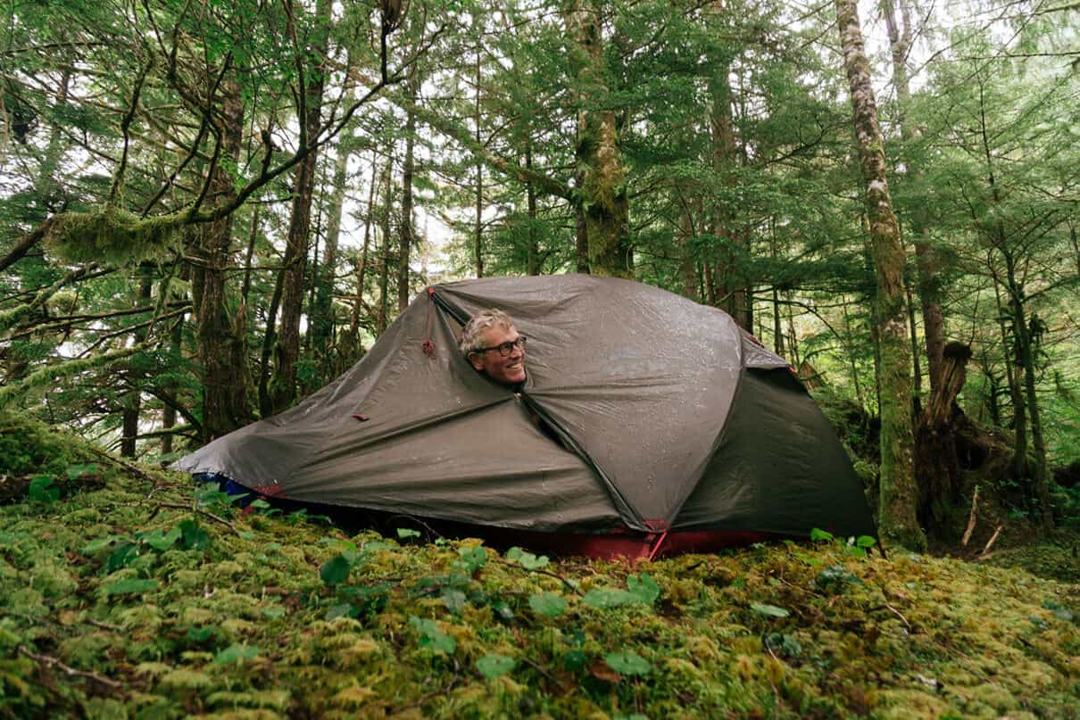 man pokes his head out of a tent in a forest campsite