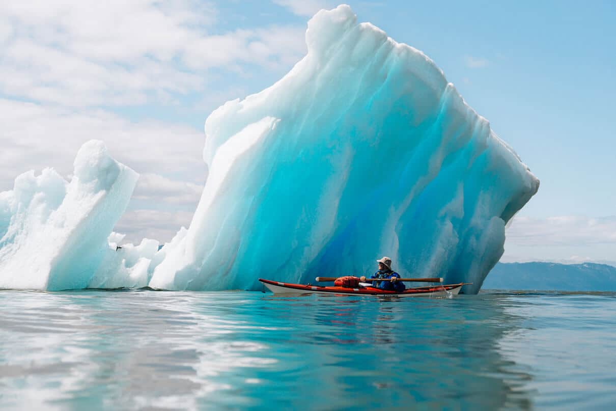man in sea kayak sits close to small iceberg with turquoise colors
