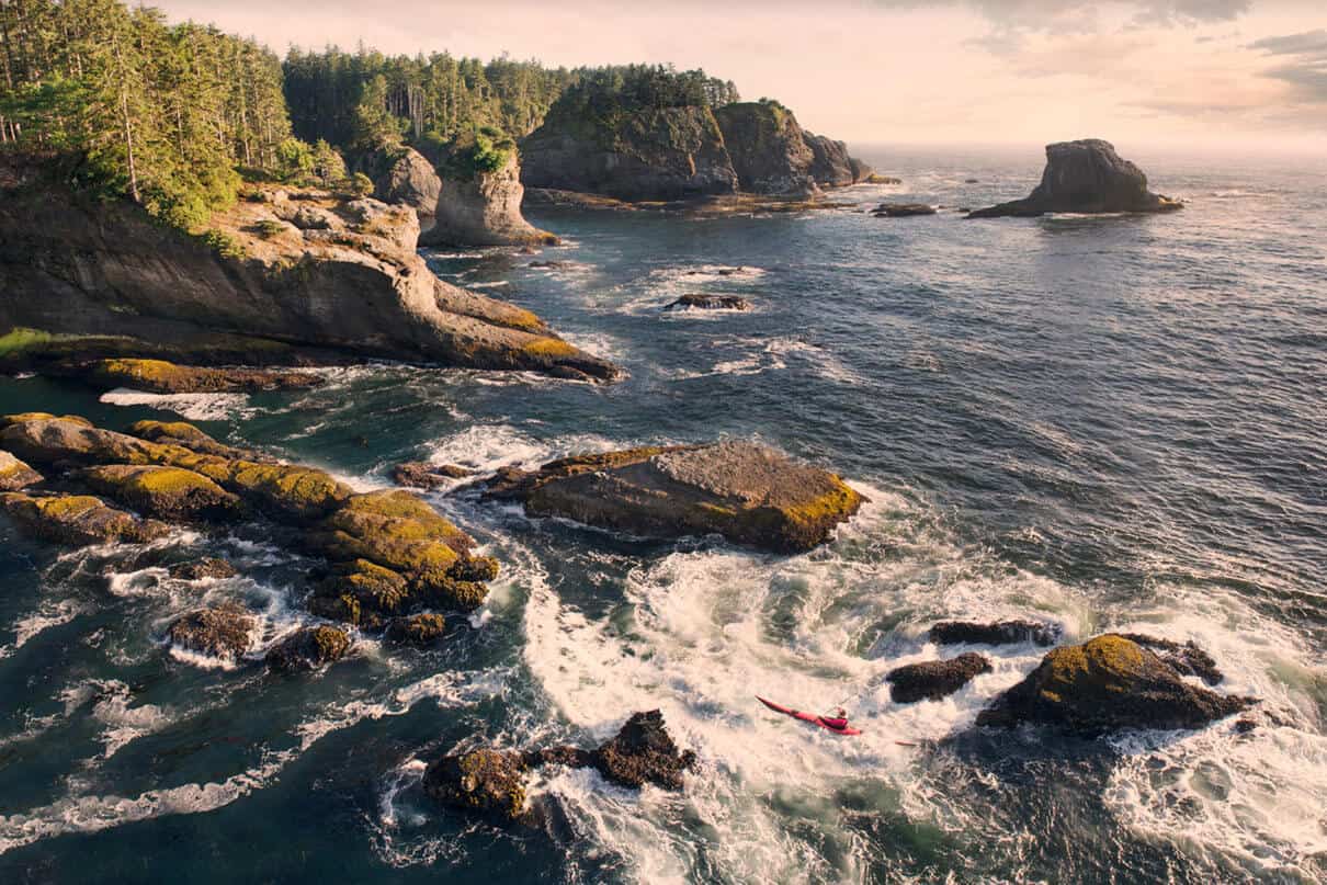 person paddles a kayak in cliffside surf on the Pacific Northwest coast
