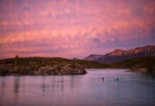 Two touring kayakers paddle in pink dawn or dusk light with mountains behind them