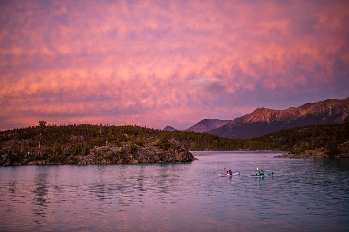 Two touring kayakers paddle in pink dawn or dusk light with mountains behind them