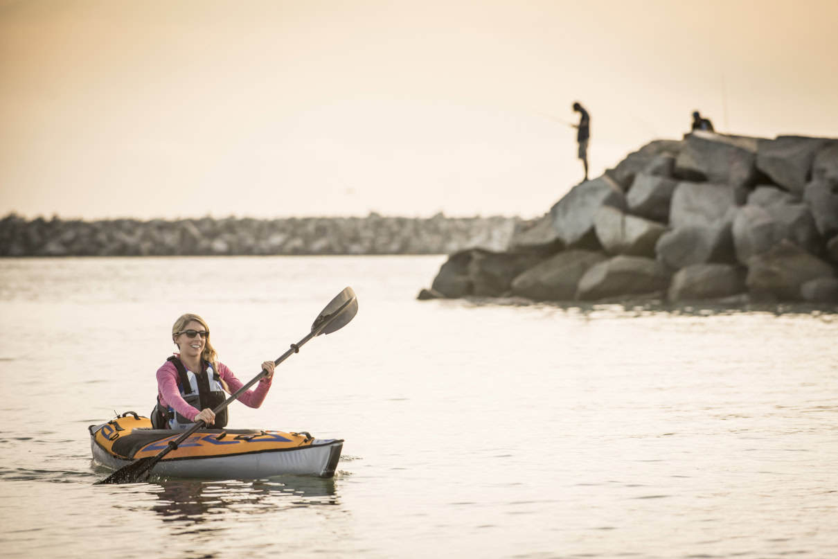 Woman paddles inflatable kayak near sea wall.