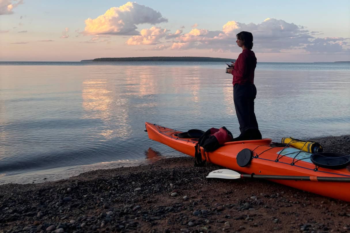Woman standing next to kayak, paddle and gear watching sunset across the water.