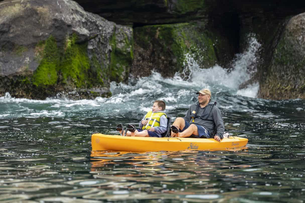 Father and son pedaling tandem kayak along a coastal cliff face.