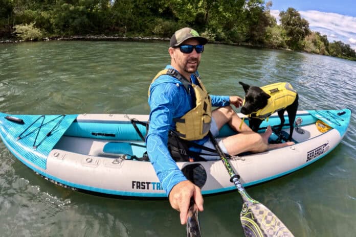 Man and dog in inflatable kayak on river.