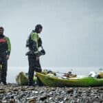 two men dressed in drysuits stand beside packed touring kayaks on a rocky shore with misty water behind them