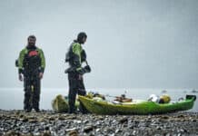 two men dressed in drysuits stand beside packed touring kayaks on a rocky shore with misty water behind them