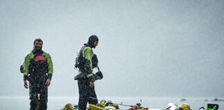 two men dressed in drysuits stand beside packed touring kayaks on a rocky shore with misty water behind them
