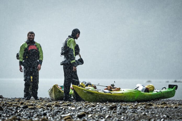 two men dressed in drysuits stand beside packed touring kayaks on a rocky shore with misty water behind them