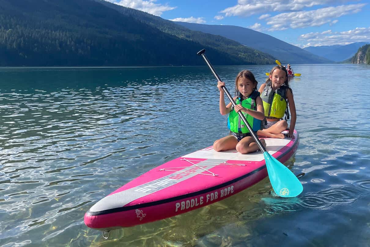 two girls kneel on an inflatable paddleboard on a western lake, the front girl is wearing a Stohlquist Drifter life jacket