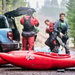 three whitewater kayakers gear up by the trunk of their vehicle with red kayaks on the ground in front