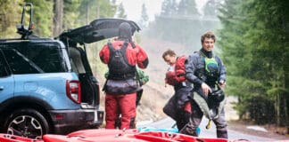 three whitewater kayakers gear up by the trunk of their vehicle with red kayaks on the ground in front