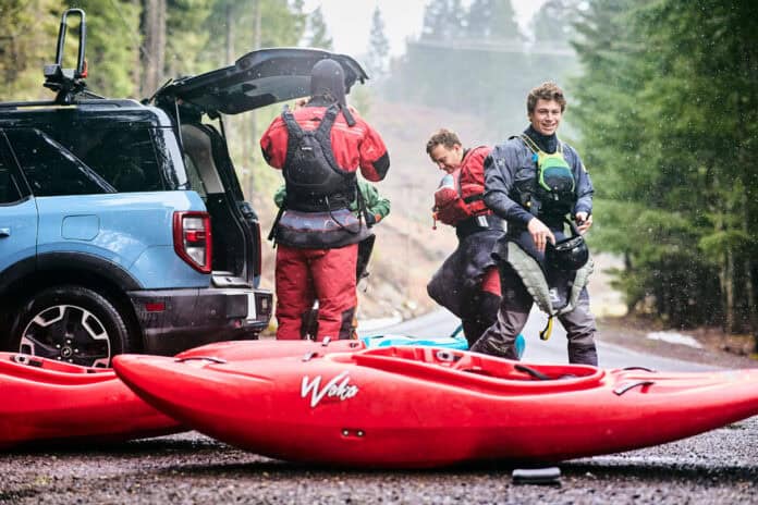 three whitewater kayakers gear up by the trunk of their vehicle with red kayaks on the ground in front