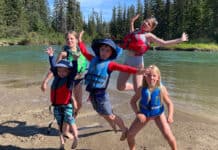a group of children and youth jump in the air beside a river while wearing a selection of life jackets