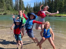 a group of children and youth jump in the air beside a river while wearing a selection of life jackets