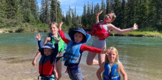Best Child And Youth Life Jackets For 2026 a group of children and youth jump in the air beside a river while wearing a selection of life jackets