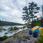 a yellow and blue tent sits on a rocky canoe campsite beside the water