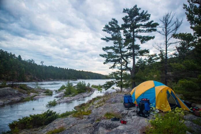 a yellow and blue tent sits on a rocky canoe campsite beside the water
