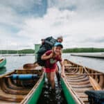 girl smiles and looks into the camera as she wades between two canoes while carrying a canoe pack at an outdoor education program, with instructor behind her