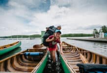 girl smiles and looks into the camera as she wades between two canoes while carrying a canoe pack at an outdoor education program, with instructor behind her