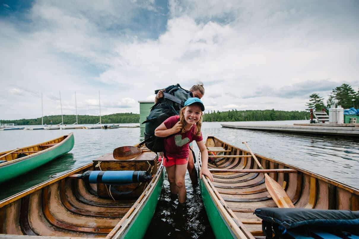 girl smiles and looks into the camera as she wades between two canoes while carrying a canoe pack at an outdoor education program, with instructor behind her