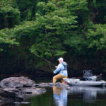 Do You Remember Your First Fishing Rod? man wades near rocks in midriver with fishing kayak beside him as he casts fly line