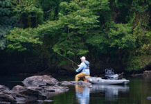 man wades near rocks in midriver with fishing kayak beside him as he casts fly line