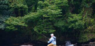 man wades near rocks in midriver with fishing kayak beside him as he casts fly line