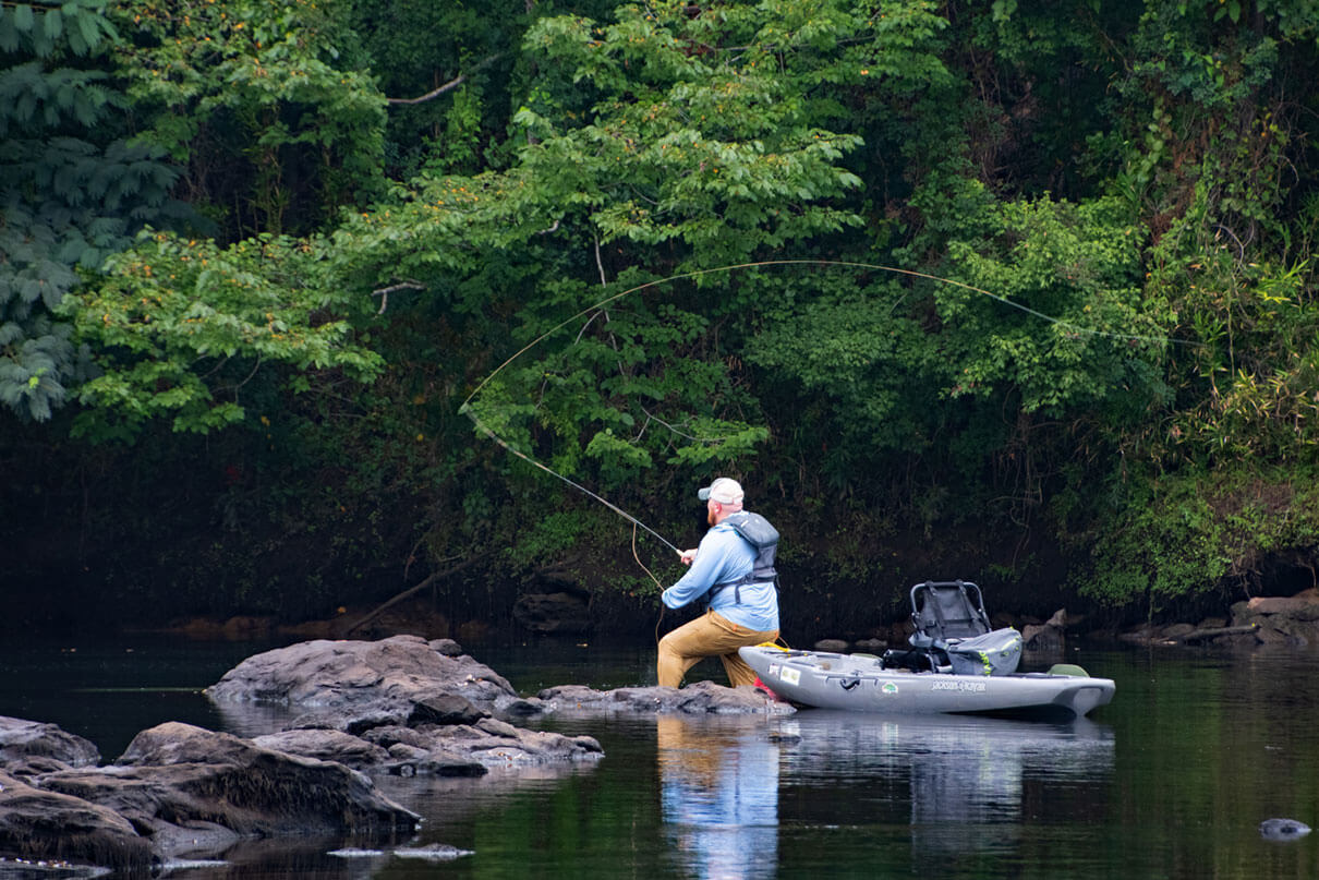man wades near rocks in midriver with fishing kayak beside him as he casts fly line