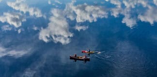 An Ode To The Company We Keep On The Water bird's eye view of a canoe and kayak paddling across still water with mountains and clouds reflected