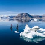 Chris Christie Dodges Icebergs In East Greenland two people paddleboard across a fjord in East Greenland, with small icebergs nearby