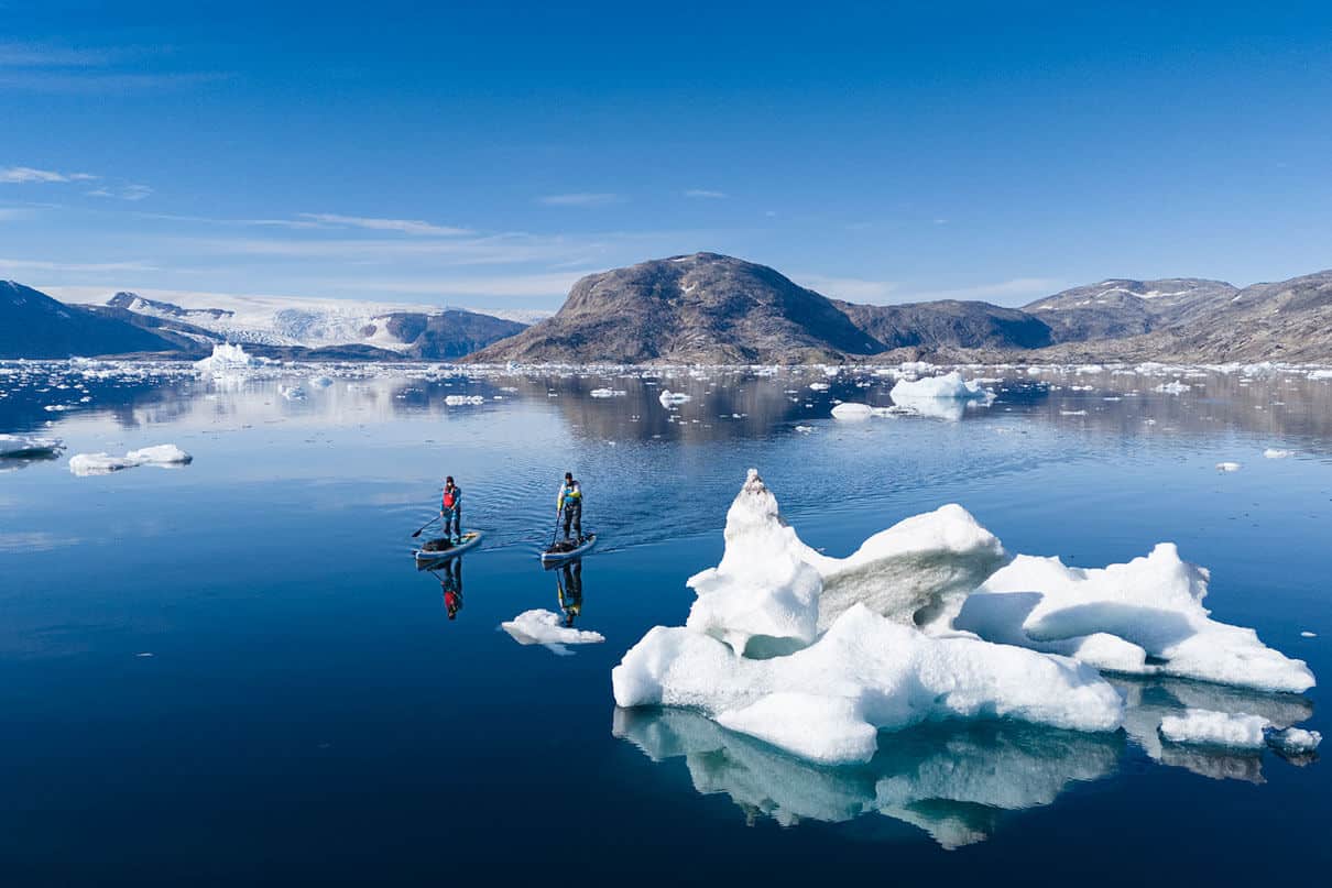 two people paddleboard across a fjord in East Greenland, with small icebergs nearby
