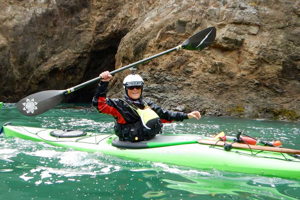 Deb Volturno on the water in green sea kayak, holding paddle aloft in front of rocky walls