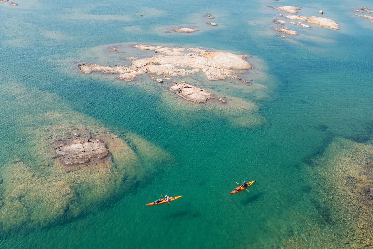 two kayakers paddle around shallow turquoise watters with rocky islands just reaching the surface