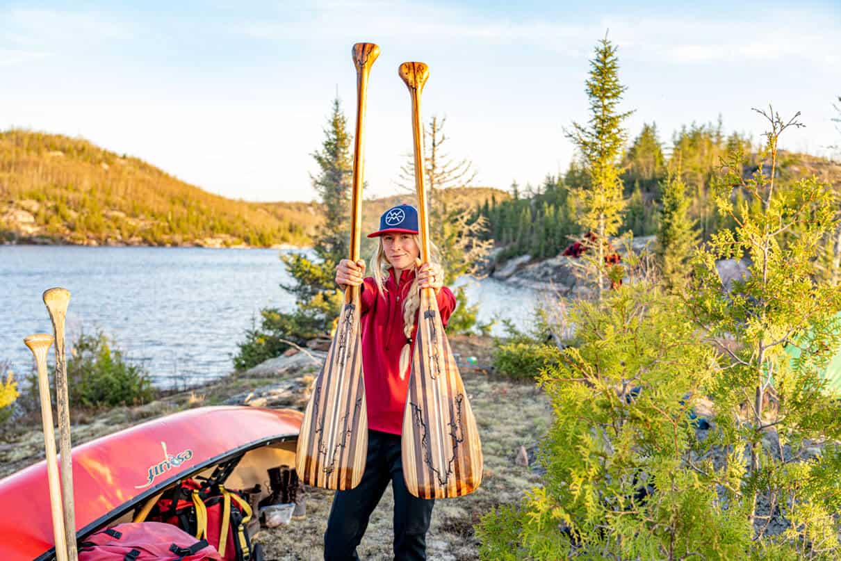 a woman holds up two wooden canoe paddles while at a northern canoe tripping campsite