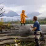 Jim Baird’s Tips For Remote Canoe Trips With Young Kids young child in yellow rainsuit stands on a tall log in a misty, mountainous wilderness setting with his dad nearby