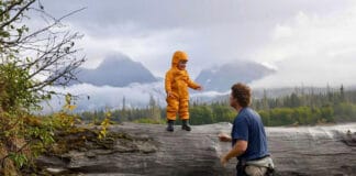 Jim Baird’s Tips For Remote Canoe Trips With Young Kids young child in yellow rainsuit stands on a tall log in a misty, mountainous wilderness setting with his dad nearby