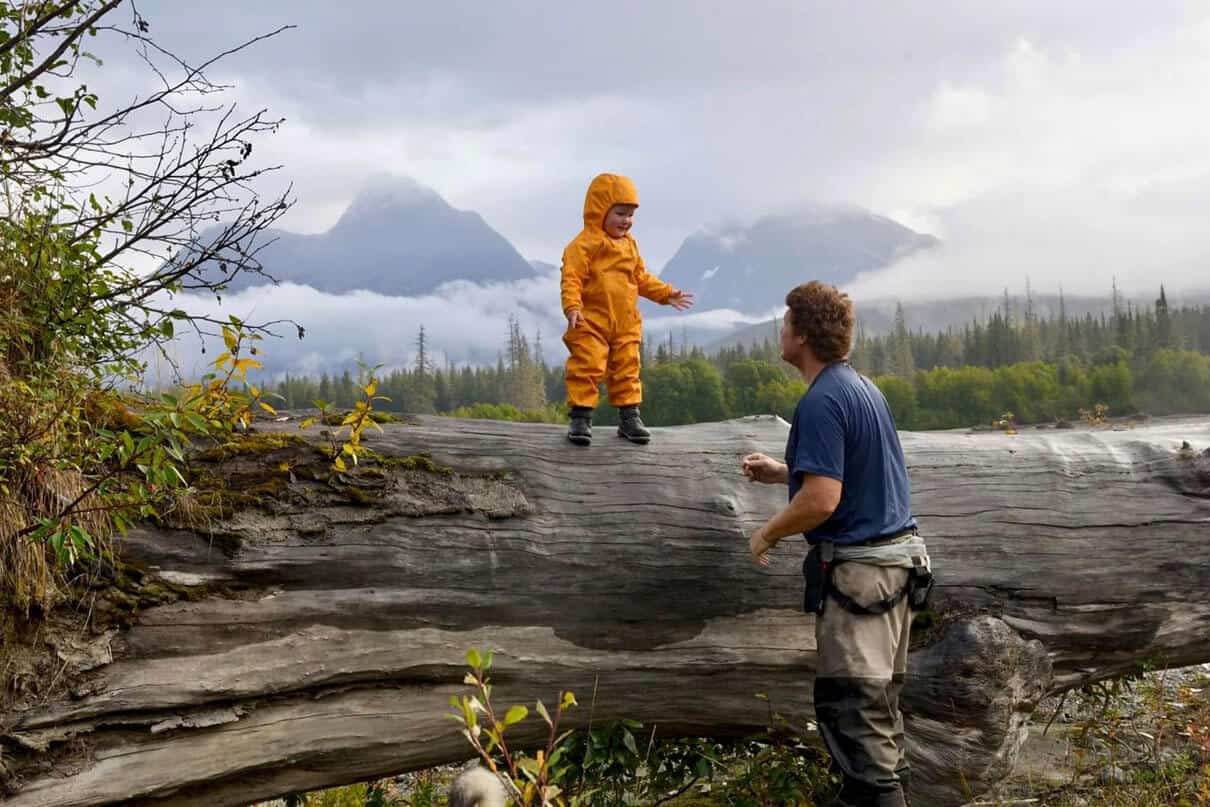 young child in yellow rainsuit stands on a tall log in a misty, mountainous wilderness setting with his dad nearby