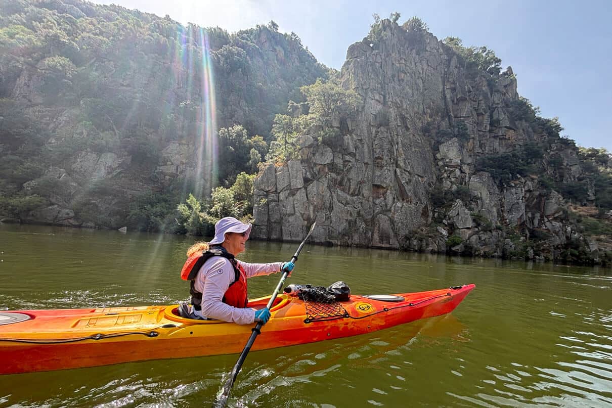 a woman paddles by rocky cliffs in the sunlight on a kayaking expedition in Portugal