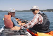 Checking Sea Time In Washington’s San Juan Islands A guide and young boy sit in kayaks side by side on the water surrounding the San Juan Islands.