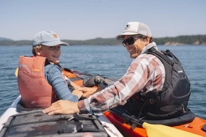 A guide and young boy sit in kayaks side by side on the water surrounding the San Juan Islands.