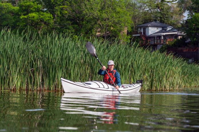 man paddles the Boreal Design Storm 17 thermoformed touring kayak in front of cattails with a house in the background