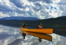 Three Pros Reimagine How To Hook New Paddlers father and child in a Clipper canoe paddling on a lake with mountains and dramatic backlit clouds behind