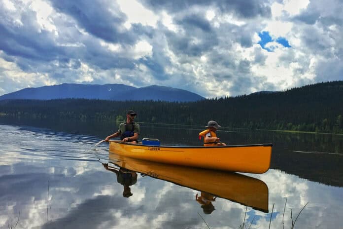 father and child in a Clipper canoe paddling on a lake with mountains and dramatic backlit clouds behind