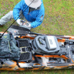 angler crouches beside fishing kayak parked on grass while working on tackle setup