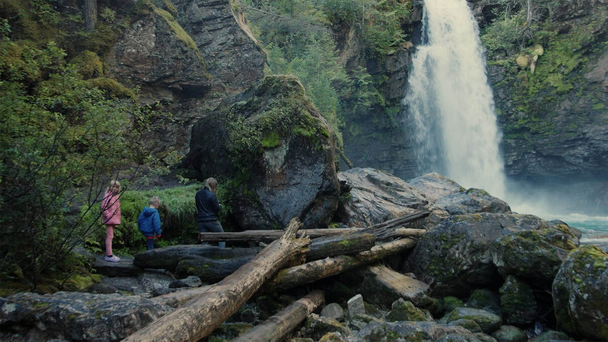 Mother and two kids stand among boulders at base of a waterfall.