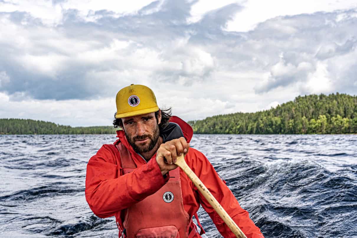 Man wearing red coat and life jacket in middle of lake and holding canoe paddle.