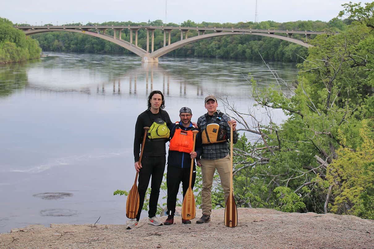 Bures meets with rescuers Jake (left) and Hunter (right) by the riverside with a bridge in the background, all three men wear PFDs and hold canoe paddles