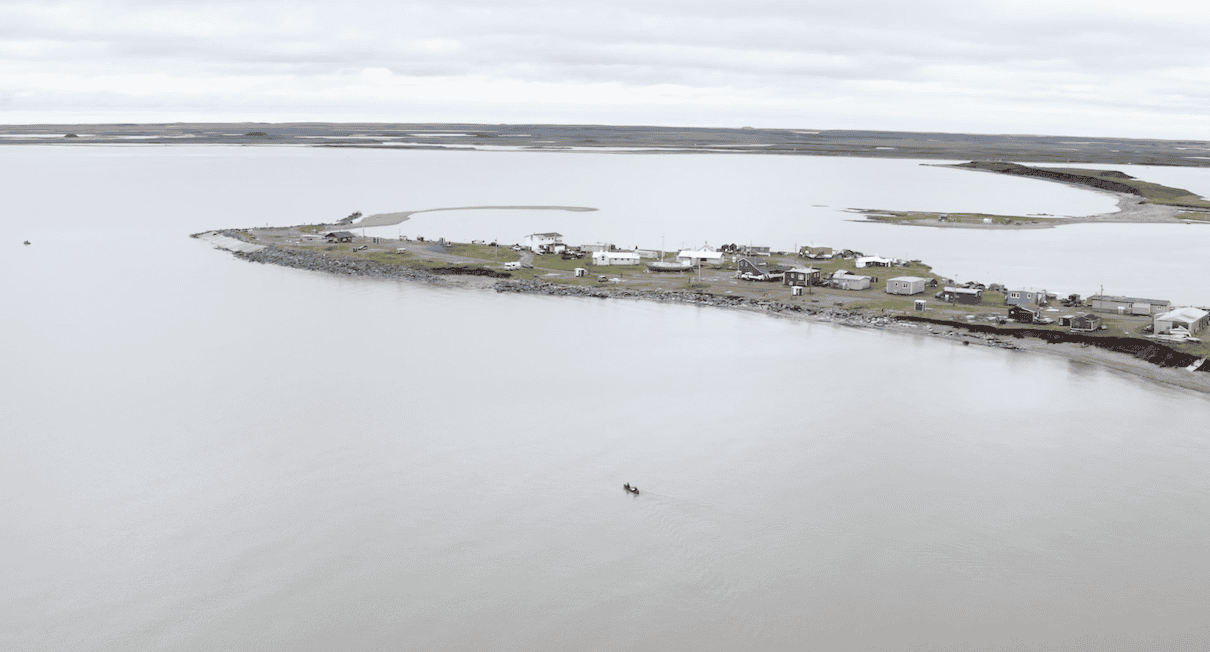 Canoe being paddled close to spit of land with houses on it sticking out into ocean.